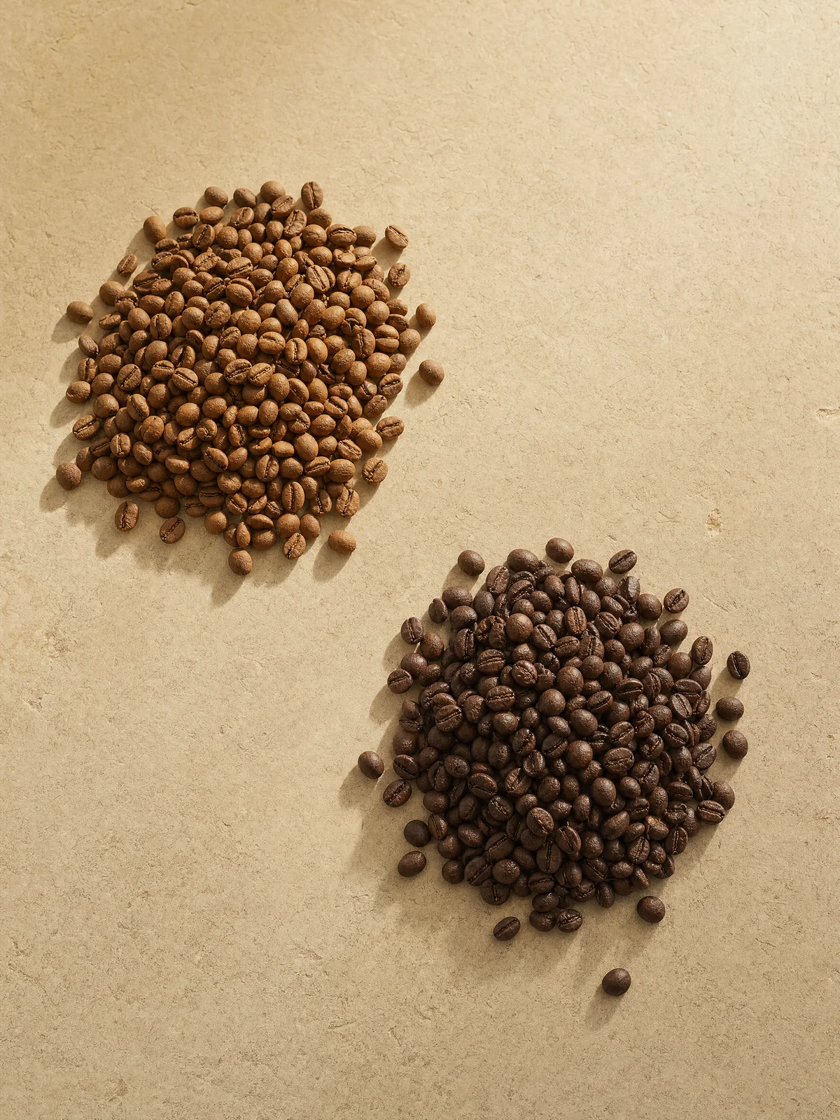 Two separate mounds of whole coffee beans, one lighter roasted and one darker roasted, on warm stone