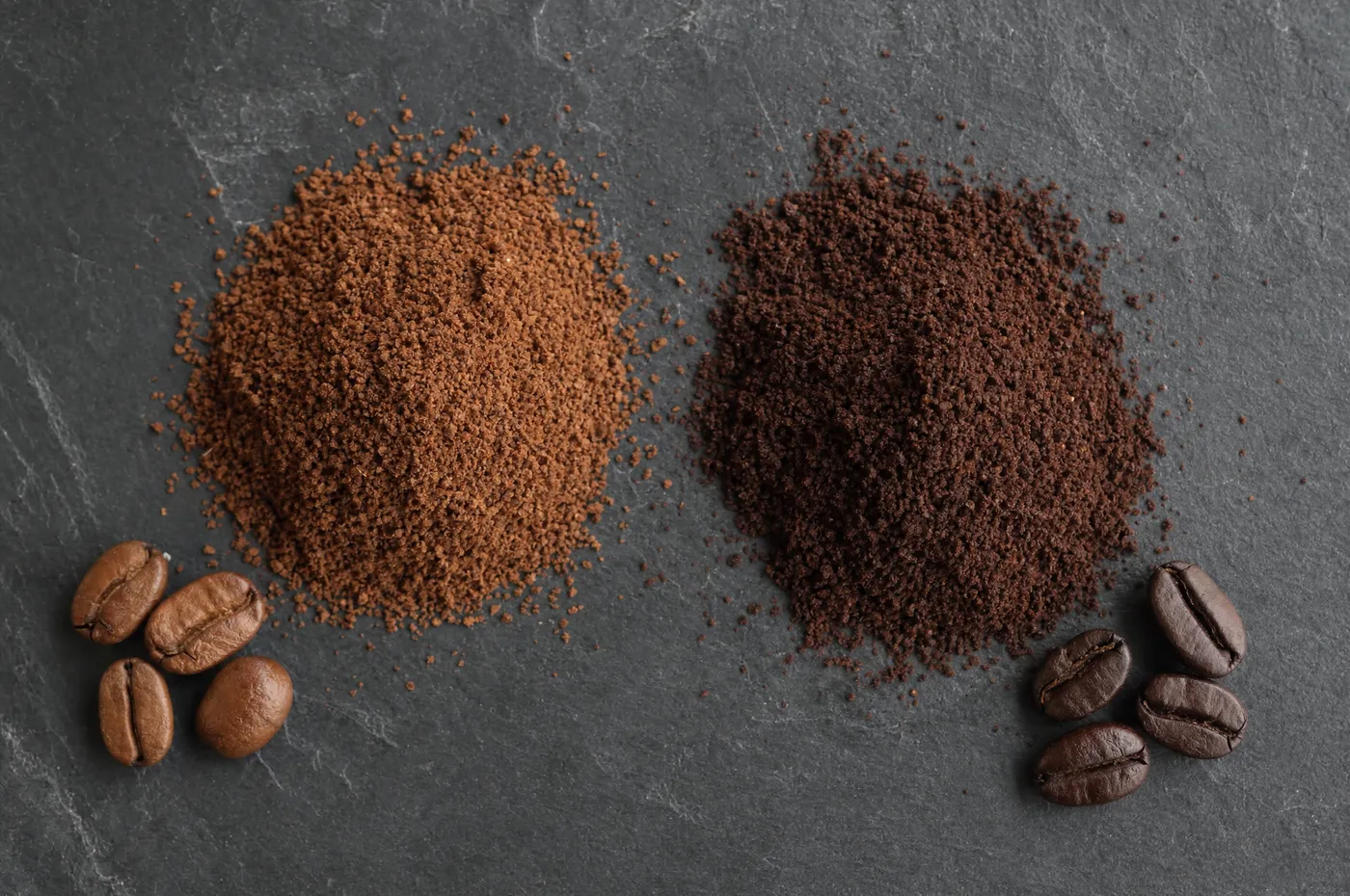 Two mounds of ground coffee on slate: light brown from a light roast, deep brown from a dark roast